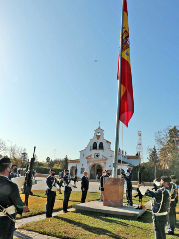 Ceremonia de izado de la bandera española en un entorno religioso, con soldados en uniforme y una iglesia al fondo.
