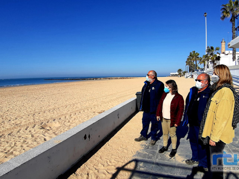 Personas con mascarillas caminando por un paseo marítimo junto a una playa desierta bajo un cielo azul claro.