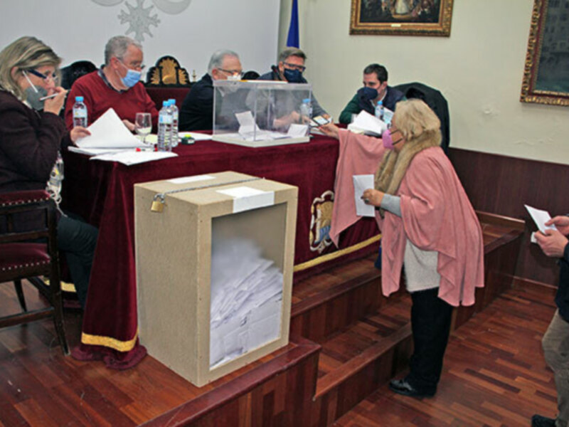 Votantes en un ambiente formal, con casillas de votación y una persona vestida con abrigo rosa.