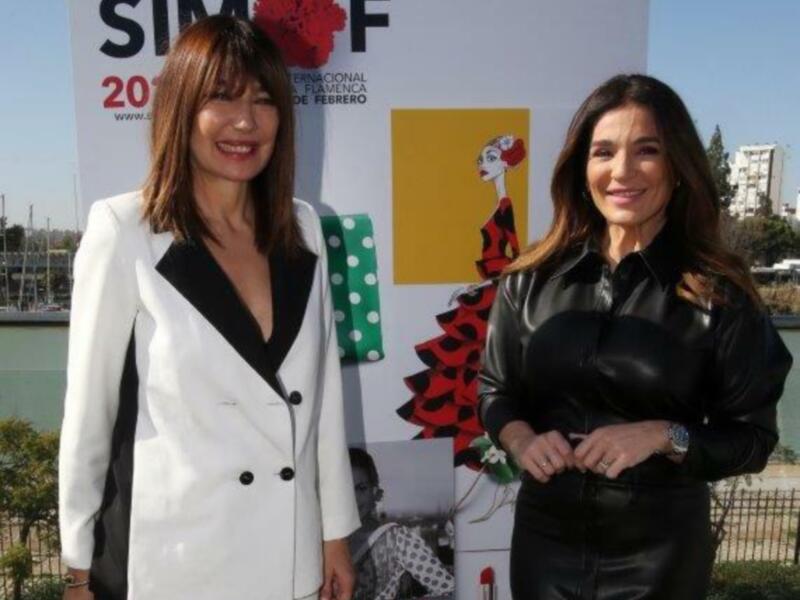 Dos mujeres posando frente a un cartel de SIMOF, evento internacional de flamenco en febrero. Una de ellas está usando un traje blanco con detalles negros y la otra una chaqueta de cuero negro con detalles plateados. Ambas sonríen para la cámara.