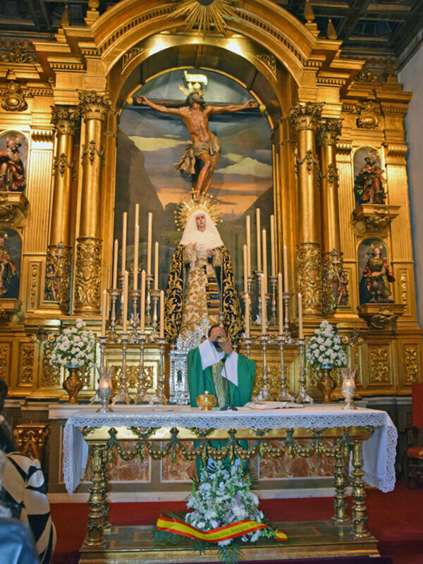Interior de una iglesia con altar dorado y imagen del Cristo crucificado, rodeado de velas y flores.