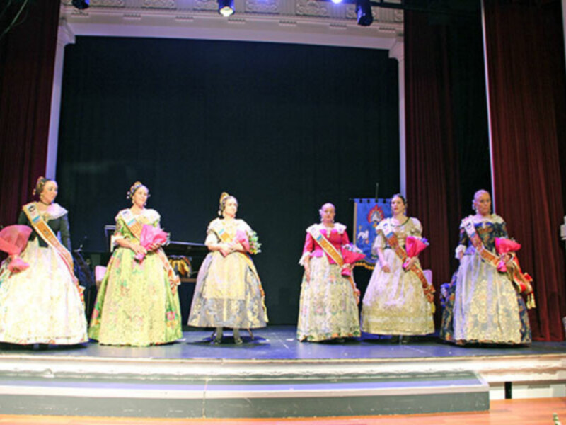 Seis mujeres vestidas con trajes tradicionales de falda y blusa, posando en una escenario con cortinas rojas y luces de teatro.
