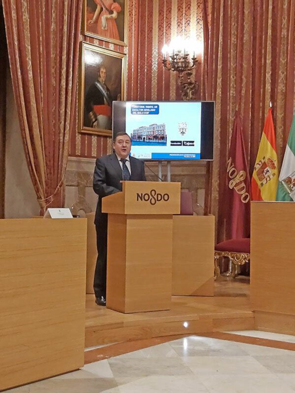 Persona hablando desde un podio en un salón formal con banderas y pantalla de presentación en el fondo.