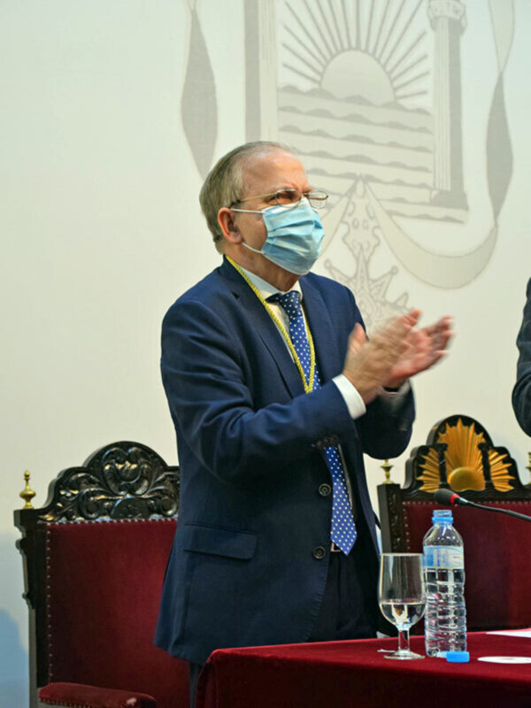 Hombre en traje con mascarilla médica aplaudiendo en un evento formal con escudo en el fondo.