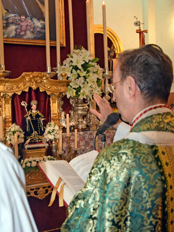 Un sacerdote en vestimenta religiosa realiza una oración en una iglesia, rodeado de velas y flores blancas, con una imagen de San Juan Evangelista en el fondo.
