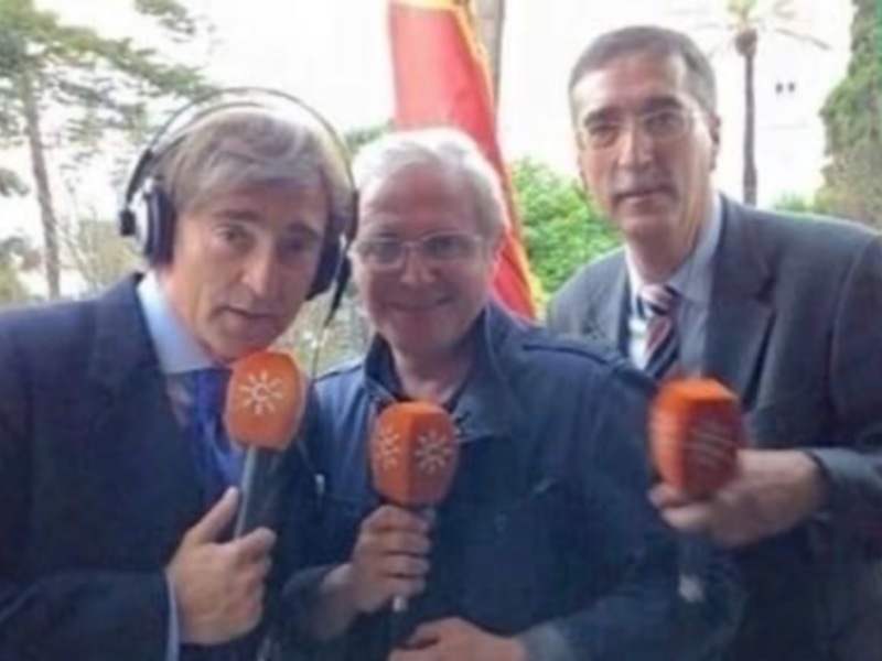 Tres personas sosteniendo micrófonos con logos de medios de comunicación, posando al aire libre con banderas en el fondo.