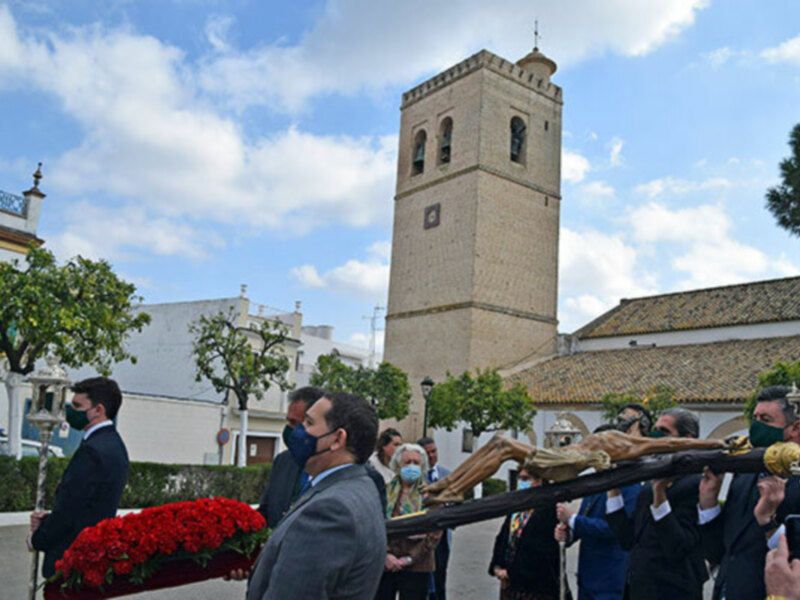 Una procesión religiosa con personas vestidas formalmente, llevando una cruz y acompañadas por una torre de mampostería en el fondo, bajo un cielo azul con nubes. La imagen transmite una atmósfera de solemnidad y devoción religiosa.