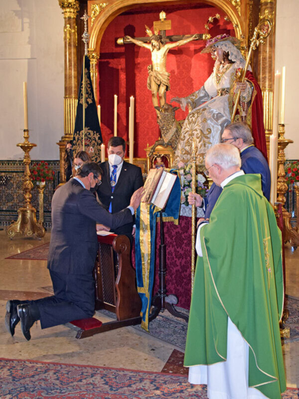 Una ceremonia religiosa en una iglesia, con un hombre en traje negro y gafas, un hombre en traje azul y corbata, y un hombre en traje verde y capa, frente a una estatua de la Virgen María en una iglesia con un altar dorado y velas encendidas.