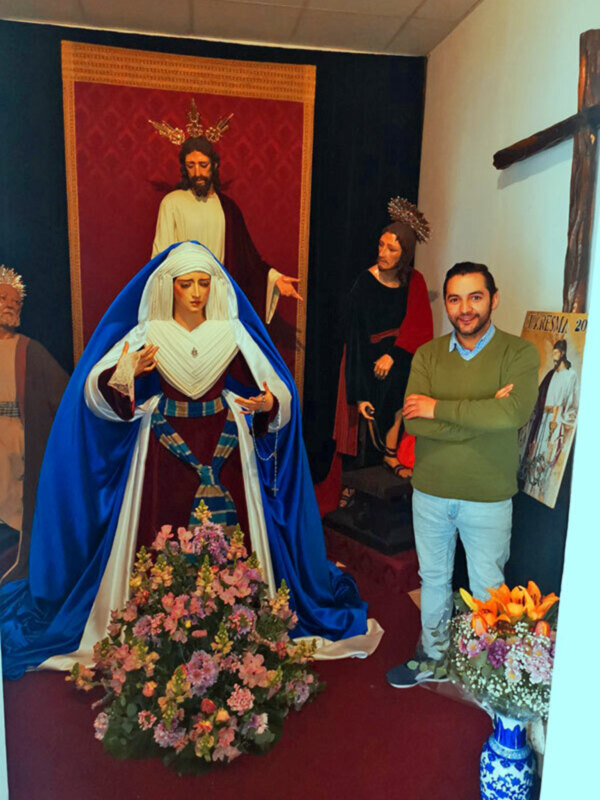 Una imagen de una figura religiosa con vestimentas tradicionales, adornada con flores, junto a una persona moderna en un entorno interno con decoración religiosa.