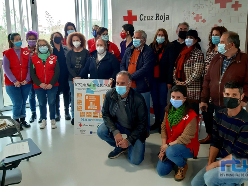 Un grupo de personas con máscaras posa frente a una bandera de Cruz Roja, sosteniendo un cartel informativo sobre un proyecto de mejora de la ciudad.