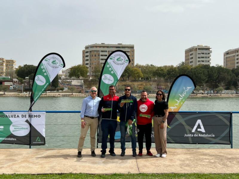 Equipo de remo de Andalucía posando junto al río Guadalquivir, con banderines de la Federación Andaluza de Remo y el lema '#RemoAndaluz'.