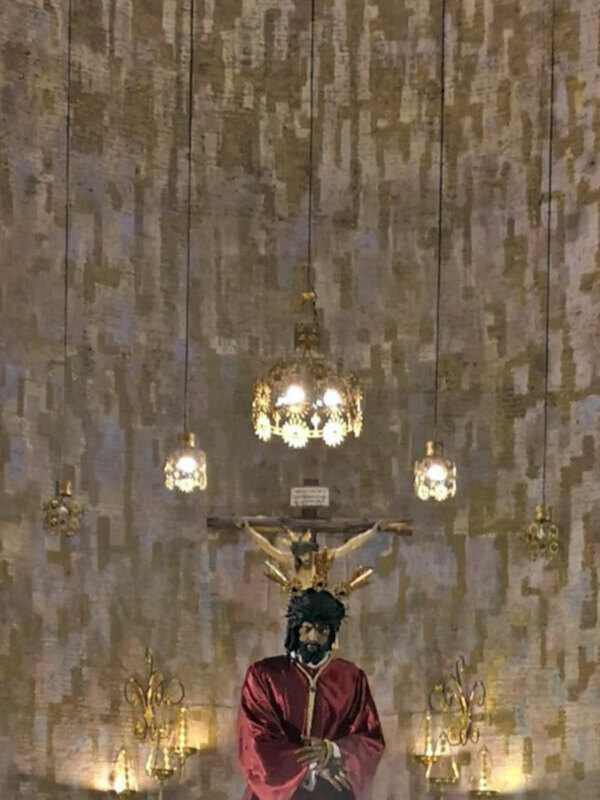 Figura religiosa en trono bajo una arquitectura de madera y lámparas colgantes en un interior de iglesia.