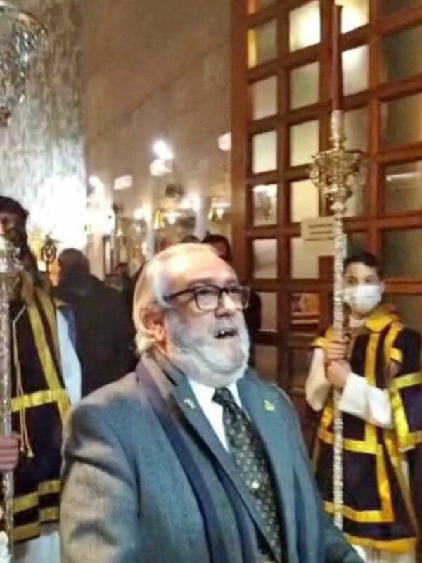 Hombre con traje y gafas hablando en un ambiente formal con decoración histórica y personas vestidas con uniformes.