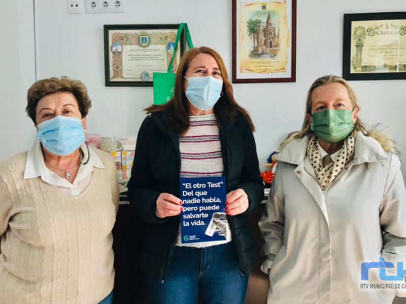 Tres mujeres con mascarillas posan con un cartel que dice 'El otro Test' en un entorno formal con certificados en la pared.