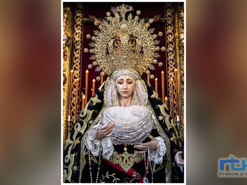 Elegante imagen de una estatua religiosa con un vestido blanco y negro, adornada con joyas y un halo dorado, rodeada de velas en un ambiente de iglesia.