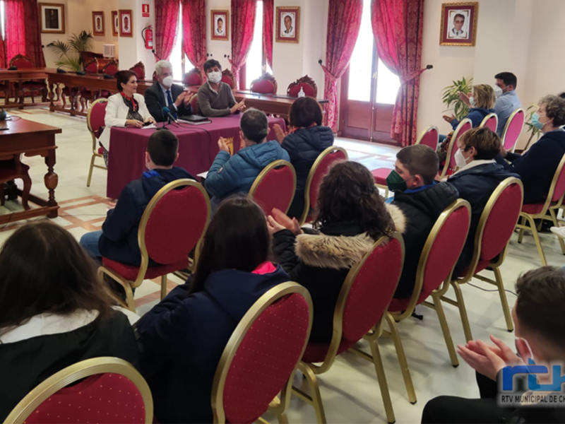 Reunión pública en un salón con personas sentadas en filas escuchando a dos individuos sentados en la mesa central.