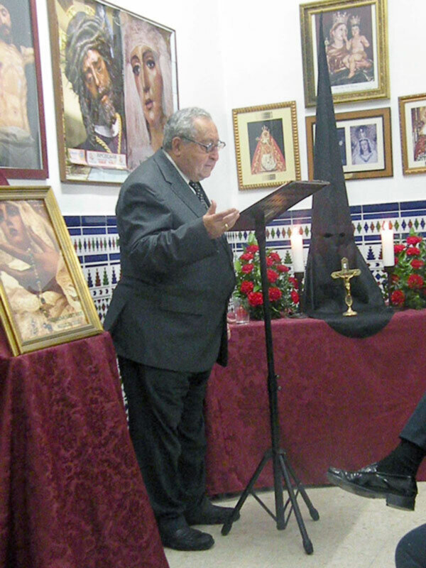 Hombre hablando en un evento religioso con imágenes de santos y flores en el fondo.