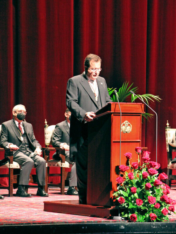 Persona hablando desde un podio en un evento formal con cortinas rojas y flores de rosas en el escenario.