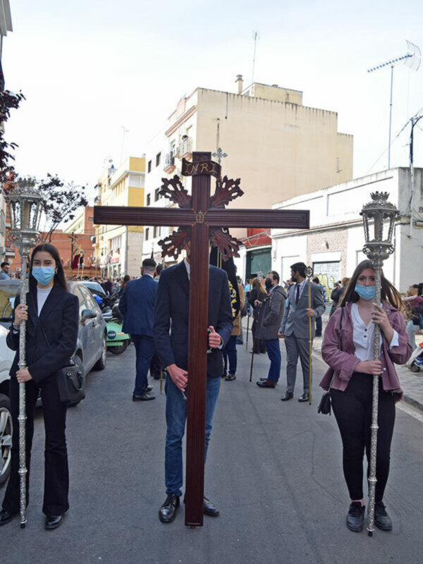 Personas con máscaras faciales y cruces durante una procesión religiosa en una calle con edificios y gente al fondo.