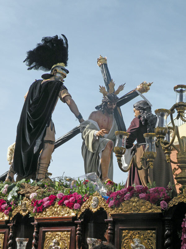 Procesión religiosa con estatuas de Jesús y soldados en un carro ornado con flores y adornos dorados.