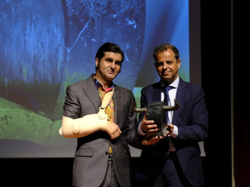 Un hombre con una prótesis de brazo y otro hombre en traje sostienen un premio en una presentación formal. El fondo muestra una imagen de la Tierra desde el espacio. La escena parece ser parte de una conferencia o evento científico.