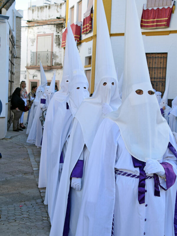Procesión de nazarenos en trajes blancos y capas, con detalles morados, en una calle urbana.
