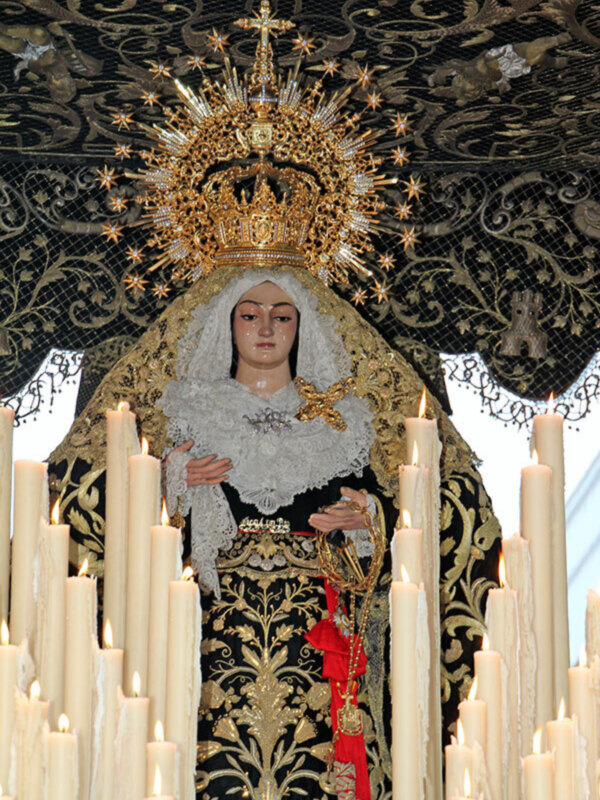 Imagen de una Virgen María con vestimentas negras y doradas, rodeada de velas y una corona de oro.