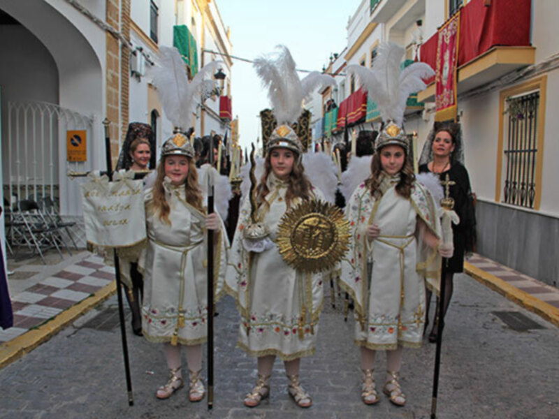 Desfile de jóvenes vestidas con trajes tradicionales en una calle decorada con banderas y palcos.