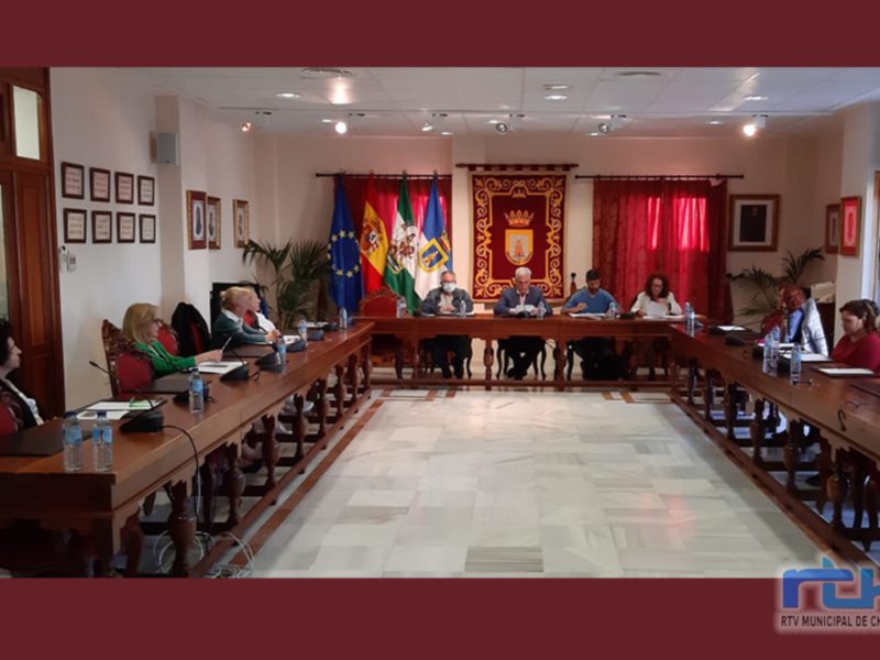 Sala de reuniones del Ayuntamiento de Chipiona con personas sentadas en mesas y banderas nacionales en el fondo.
