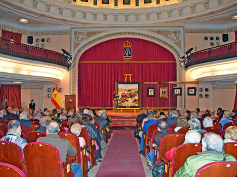 Interior of a grand auditorium with red curtains, ornate decorations, and a seated audience facing a stage with a Spanish flag and emblem.