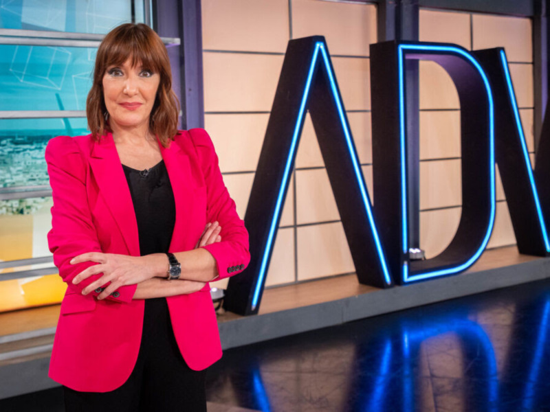 Presentadora en un estudio de televisión con el logo 'ADV' iluminado al fondo.