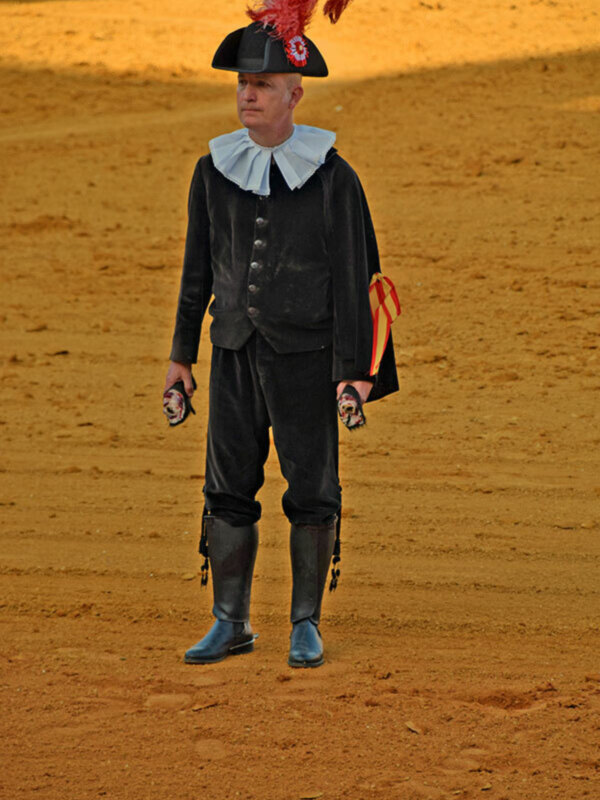 Persona vestida con traje tradicional español, incluyendo un sombrero con pluma roja, en un entorno de arena.