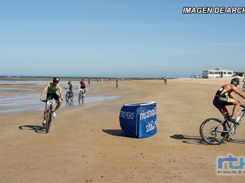 Ciclistas en una carrera por la playa, con el mar y el cielo azul al fondo.
