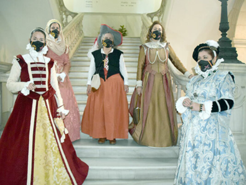 Recreación histórica de vestimenta renacentista con máscaras protectoras en un escenario interior con escaleras.