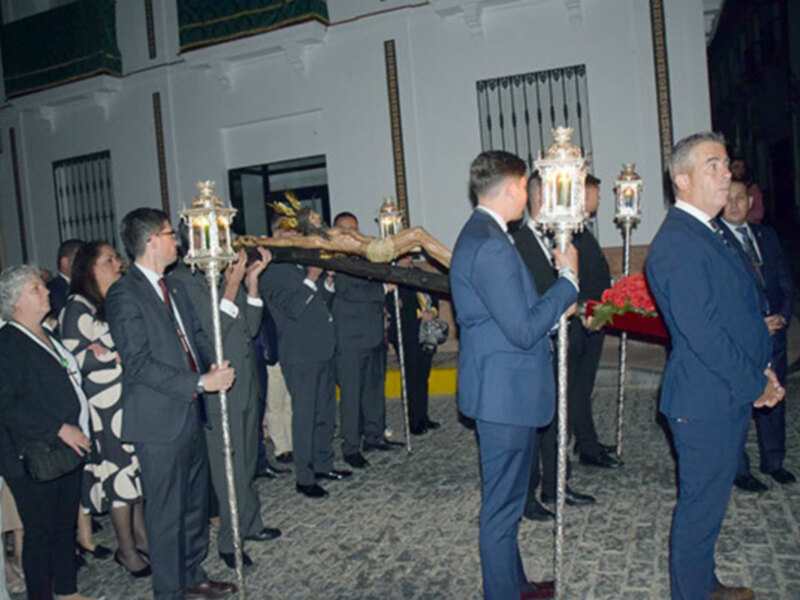 Un grupo de personas en trajes formales sostienen un bastón ceremonial en una ceremonia nocturna. La escena parece ser un evento formal, posiblemente una boda o una ceremonia religiosa, con luces y decoración en el fondo.