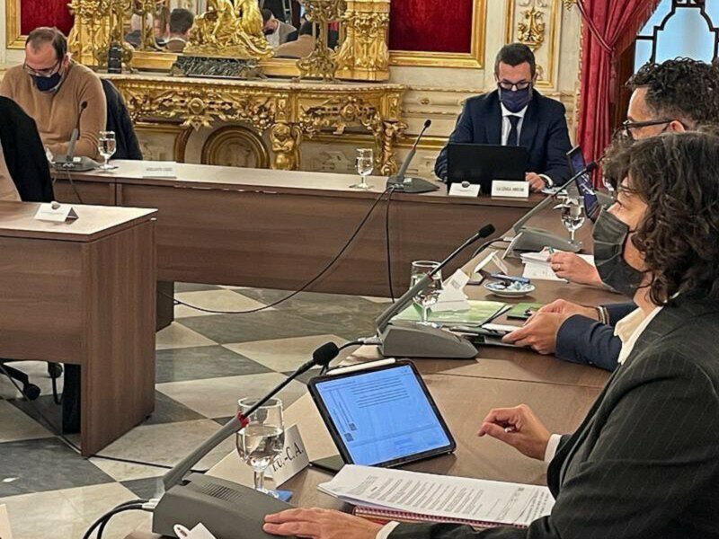 Reunión de gobierno en un salón decorado con dorado, participantes con máscaras y documentos en mano.
