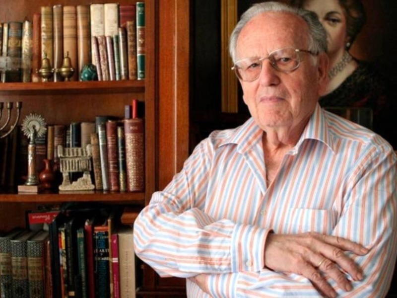Un hombre mayor con gafas y camisa a rayas, con los brazos cruzados, frente a una estantería llena de libros y objetos decorativos.