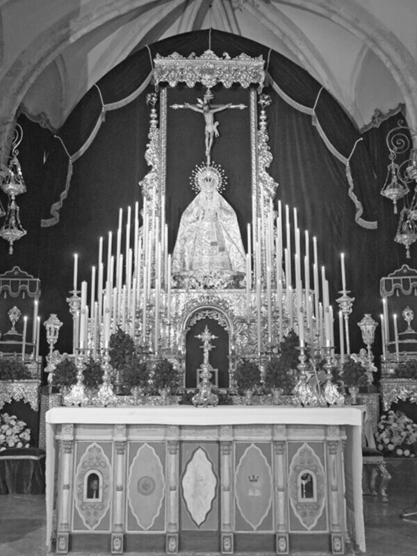 Altar con una imagen de la Virgen María en el centro, rodeada de velas y flores, con un fondo oscuro y detalles dorados.