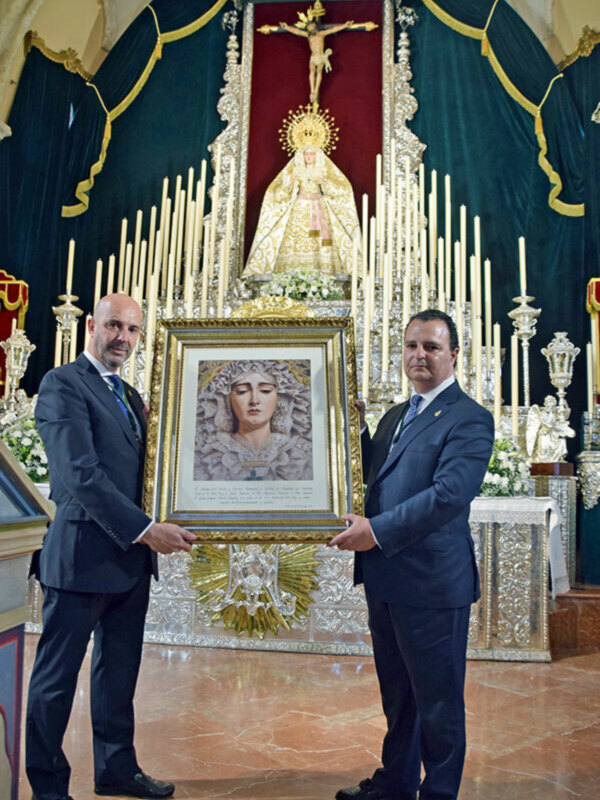 Dos hombres en traje sostienen un cuadro en un altar con la Virgen de la Amargura y el Cristo de la Buena Muerte.