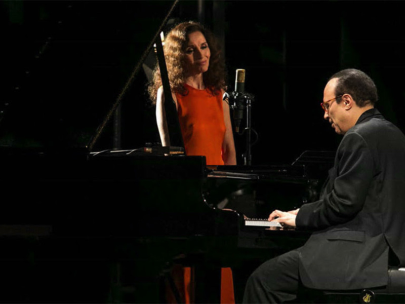 Un pianista en un concierto, con una mujer en el fondo, tocando el piano en un escenario iluminado.