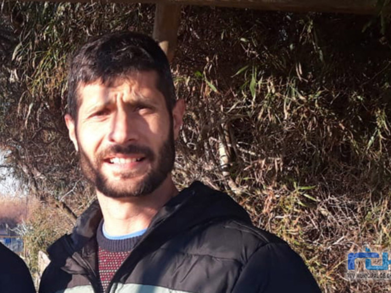 Hombre con barba y bigote, vistiendo una chaqueta negra, posando al aire libre con un fondo de vegetación densa.