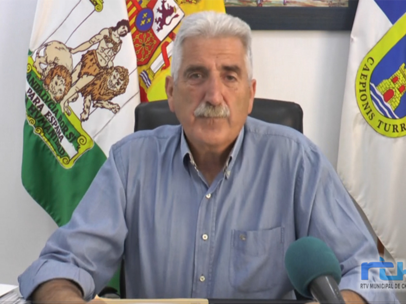Persona hablando durante una transmisión de RTV Municipal de Chipiona, con banderas y escudos de Chipiona y España en el fondo.