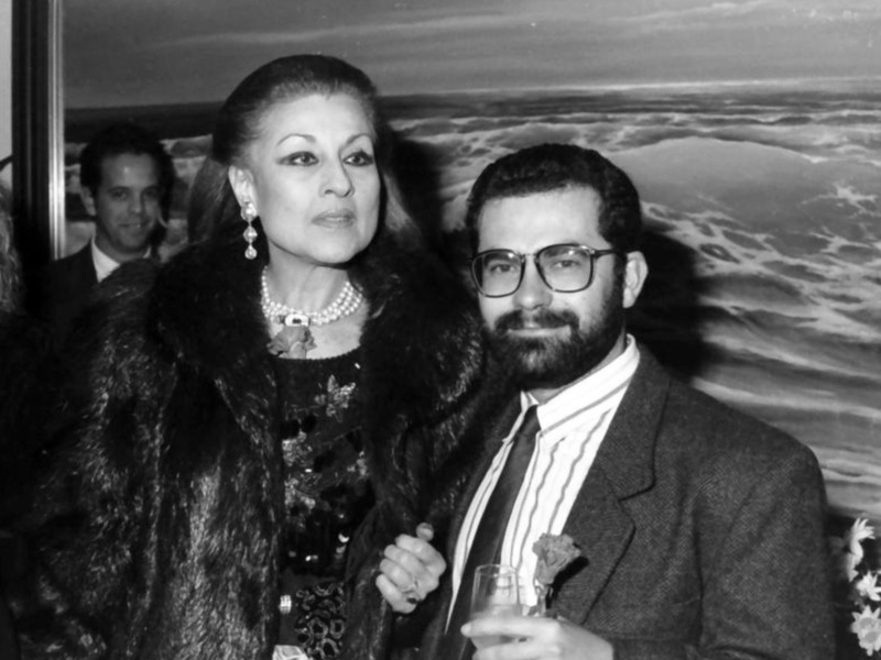 Persona elegante en abrigo de piel con perlas, posando con hombre en traje y gafas en evento formal.