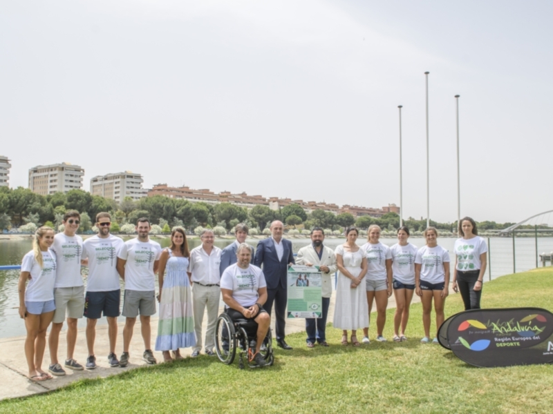 Equipo de remo de la Selección de Andalucía posando junto a un lago con edificios y banderas en el fondo.