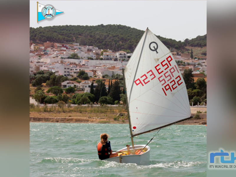 Vela de regata con el número 923 ESP 55PI 1922 navegando en el agua con un paisaje urbano en el fondo.