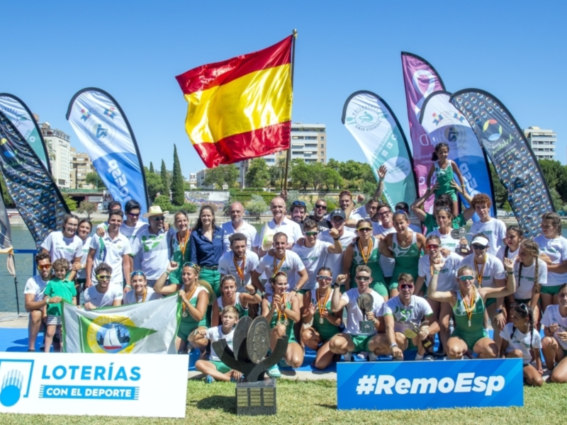 Equipo español de remo celebrando victoria con bandera española y trofeos en un evento deportivo al aire libre.