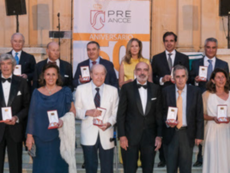 Una foto de grupo de personas en trajes formales, posando para una foto en un evento de premios, con un fondo que incluye un cartel con texto en español.