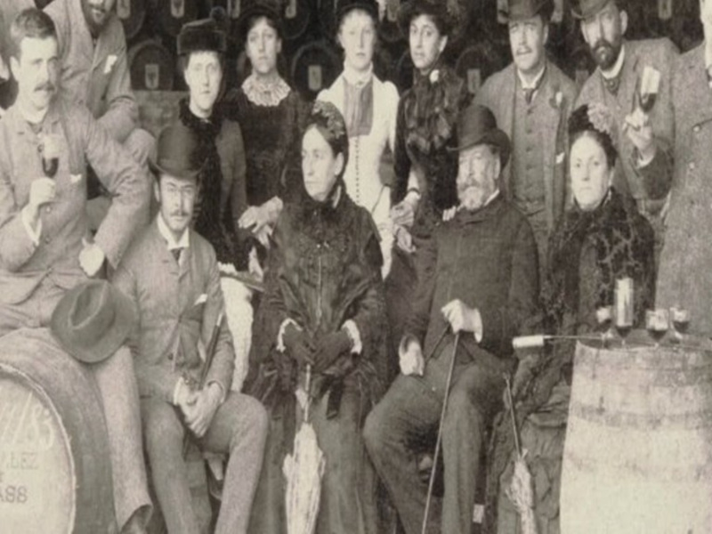 Grupo de personas vestidas con trajes y sombreros de época posando en un entorno con barriles de vino, probablemente en una bodega o fábrica de vino.