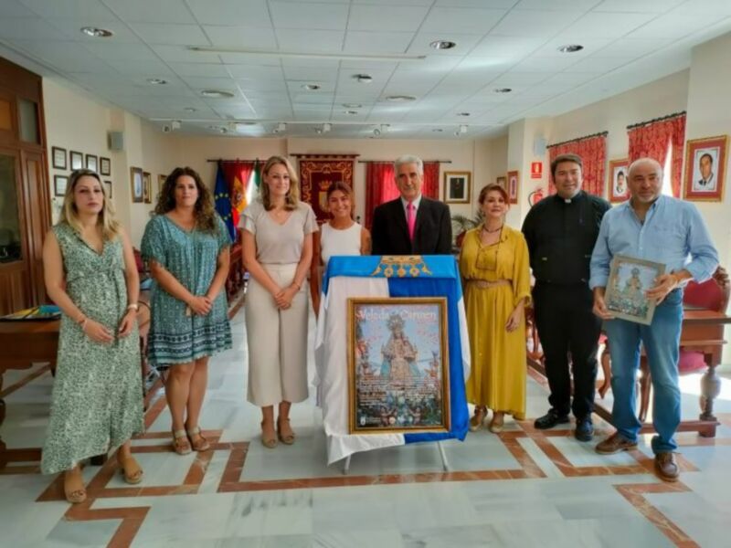 Una reunión formal en un salón con una mesa y sillas, donde se presentan varias personas vestidas formalmente. En el centro, se puede ver un cartel con una imagen religiosa y un documento. La atmósfera es formal y profesional.