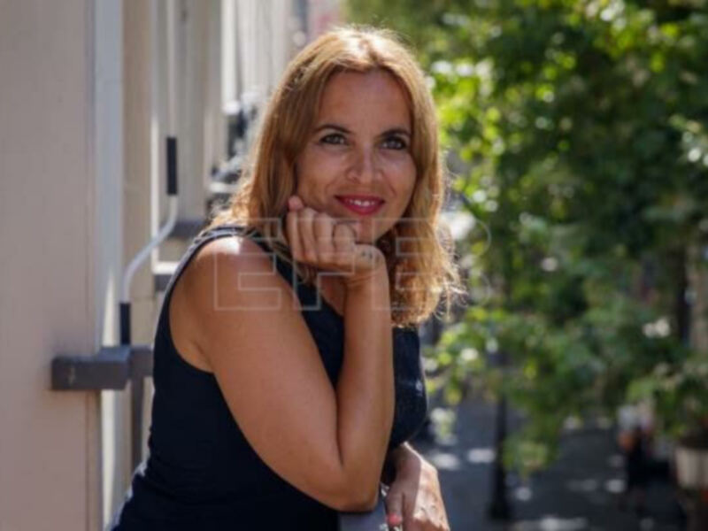 Mujer sonriente apoyada en el balcón, con un fondo de calle adoquinada y árboles verdes.